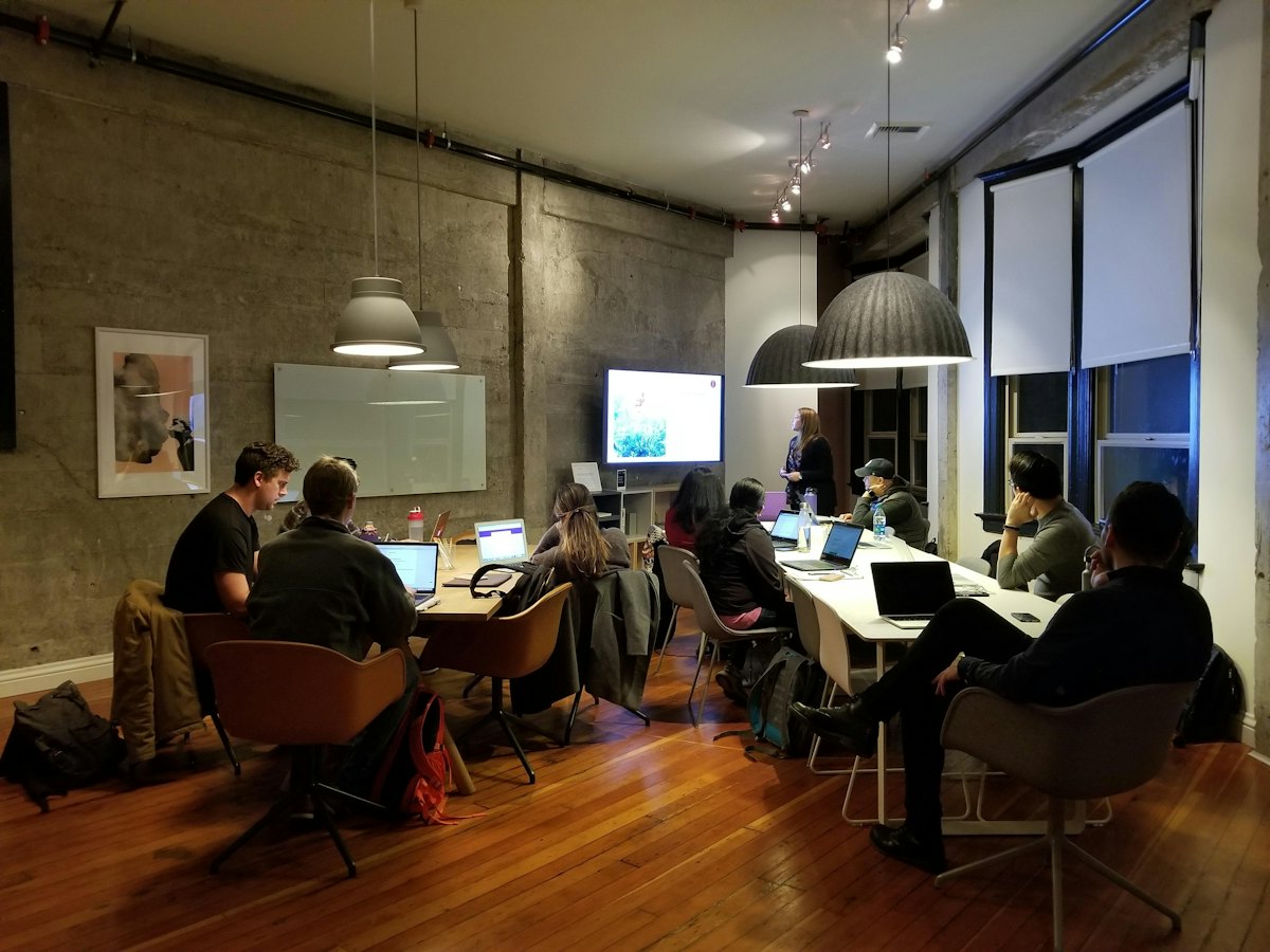 A group of people working on laptops at long tables in a warm-lit loft