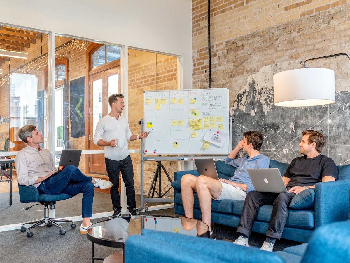 A small team on sofas with laptops, one person presenting at a whiteboard