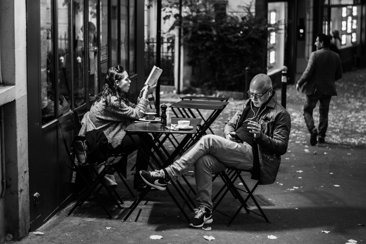 Two people at a street cafe table, one reading, one on a phone
